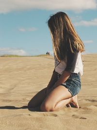 Side view of young woman kneeling on sand at beach against sky during sunny day