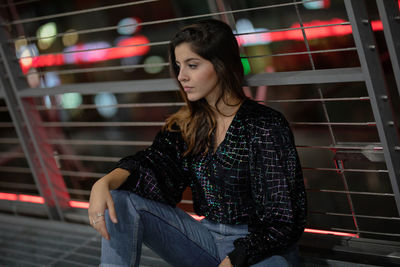 Young woman looking away while sitting against railing at night