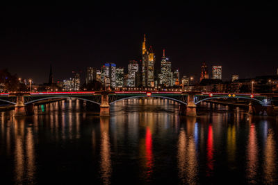 Illuminated bridge over river by buildings against sky at night