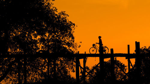 Woman with bicycle on footbridge standing by silhouette tree against clear sky during sunset