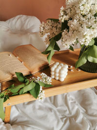 Morning in bed with book, bubble cube soy candle and bunch of white lilac on folding wooden table.