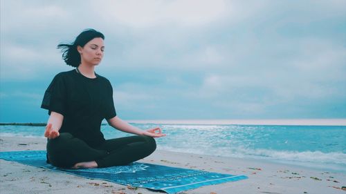 Woman meditating while sitting on shore at beach against sky