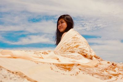 Young woman wrapped in blanket on sand at beach against sky