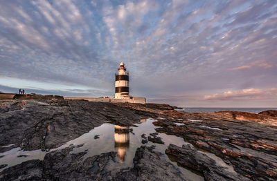 Lighthouse by sea against cloudy sky during sunset