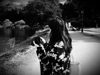 Side view of woman holding camera on street by lake during sunny day