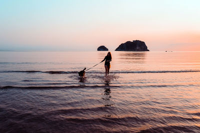 Silhouette mid adult woman with dog standing at beach against sky during sunset