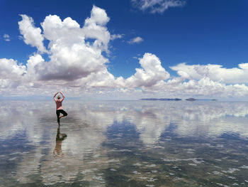 Rear view of woman practicing yoga in sea against cloudy sky
