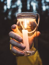 Close-up of hand holding burning candle in glass jar at forest during dusk