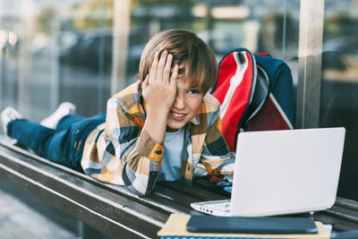 Boy using laptop while lying on bench by modern building