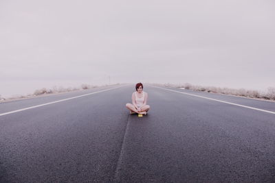 Full length of woman sitting on country road against sky during foggy weather