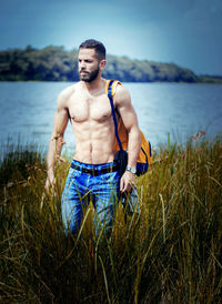 Shirtless young man looking away while standing on land by lake