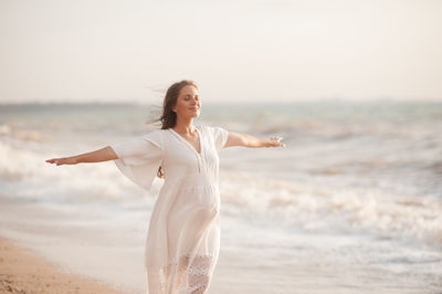 Happy pregnant woman wear white dress feeling free posing over sea background at beach. motherhood. 