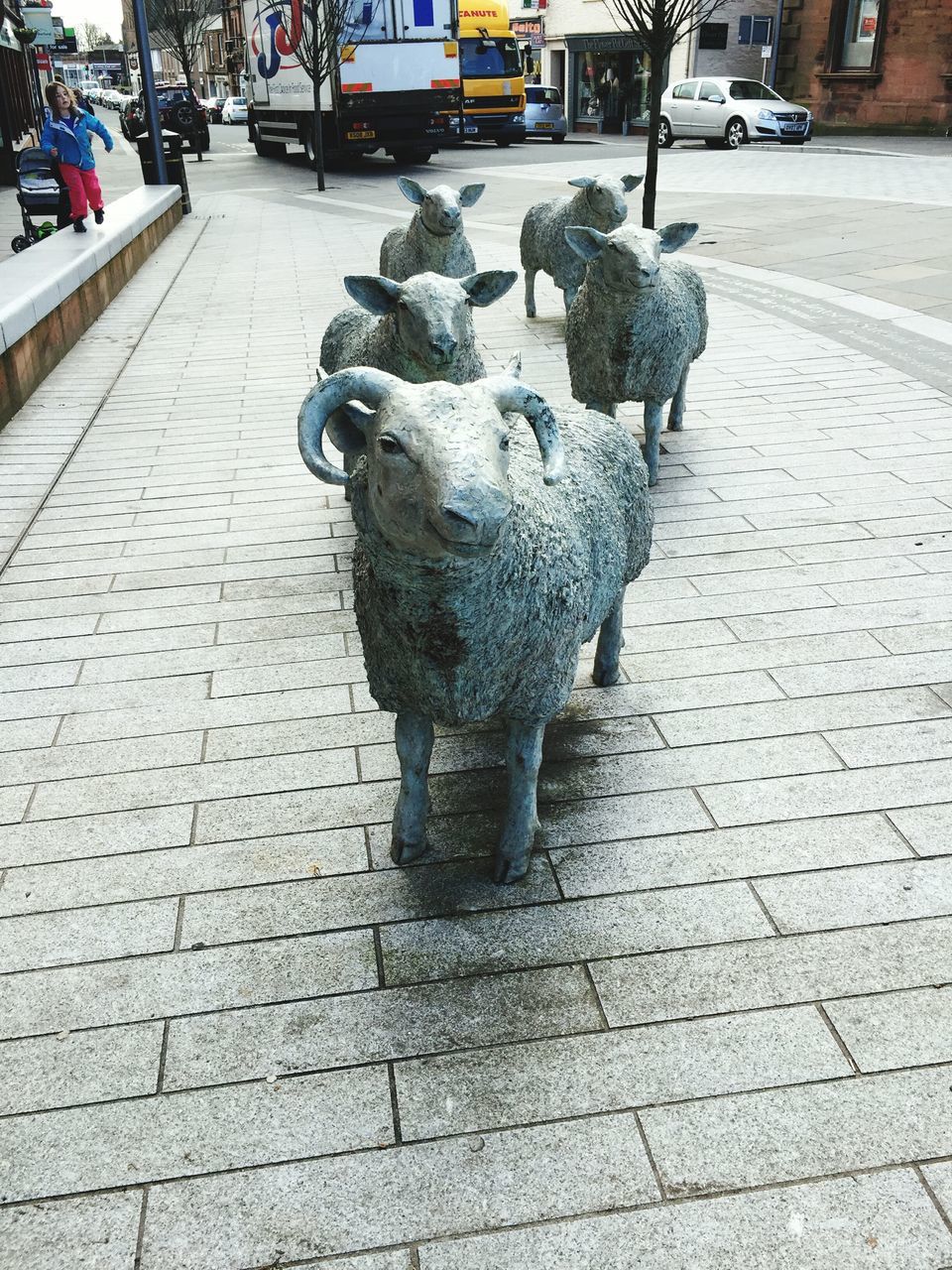 street, sidewalk, cobblestone, incidental people, paving stone, road, transportation, city, footpath, pavement, outdoors, art and craft, human representation, car, art, day, building exterior, sculpture, statue, creativity