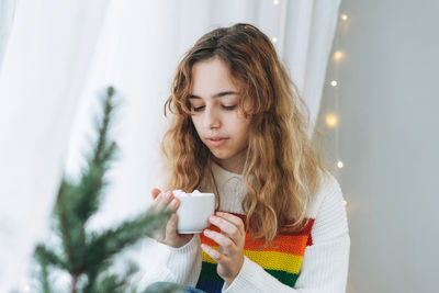 Beautiful teenager girl with cup of cacao with marshmallow sitting on windowsill in cozy room