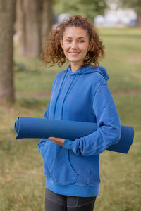 Woman in a blue hoodie with a blue yoga mat chooses a place to do sports and meditation