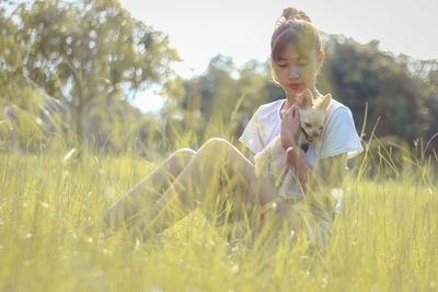 Young woman carrying dog while sitting on grassy field during sunny day