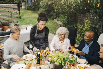 Woman serving grilled meal to senior friends sitting at dining table during dinner party in back yard