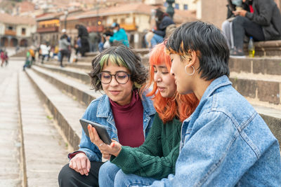 Delighted positive female friends sitting on steps of staircase in city of cusco and looking video on tablet