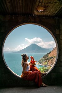 Side view of young woman sitting in window with scenic view of volcano in guatemala 