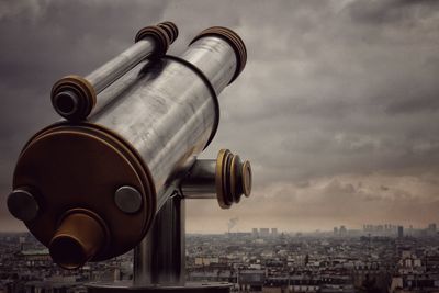 Coin-operated binoculars by cityscape against cloudy sky