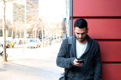 Front view of a bearded man using phone leaning on office building wall