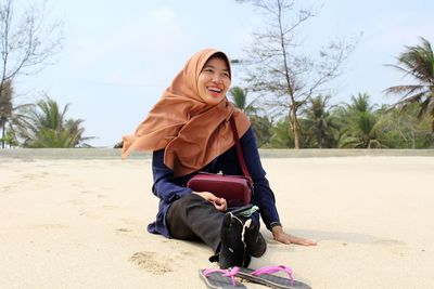 Full length of smiling young woman wearing hijab sitting at beach