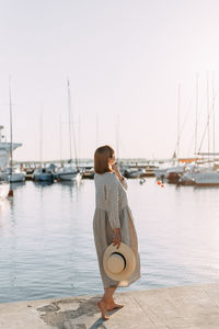 Happy young woman in dress and hat walks on the beach near the boats