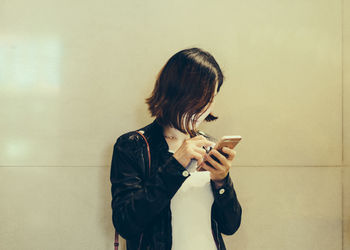 Young woman using phone while standing against wall