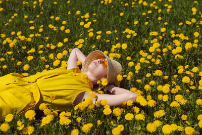 Woman in a yellow dress, hat and yellow glasses lies on a field of dandelions in summer