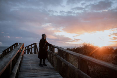 Side view of elderly female traveler in casual clothes standing on wooden pier on sandy beach and enjoying seascape at sunset