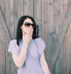 Young woman wearing sunglasses smoking standing against wood