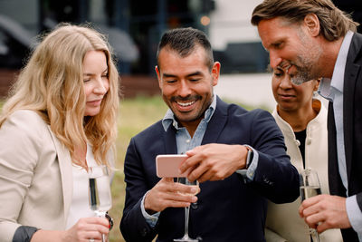 Smiling businessman showing mobile phone to male and female colleagues while holding champagne flute