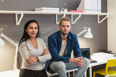 Portrait of confident business colleagues sitting on desk against wall at creative office