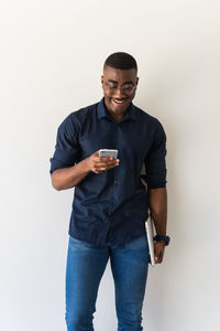 African american adult male in stylish outfit and eyeglasses standing with netbook in hands while using cellphone on white background