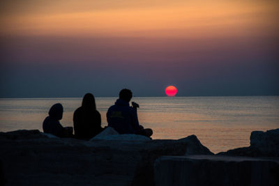 Friends sitting on sea shore against sky during sunset