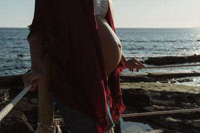 Cropped unrecognizable pregnant female in hat while standing near fence against sea and blue sky while resting on coast on summer day