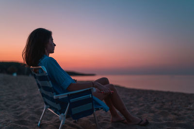 Woman sitting on beach by sea against sky during sunset