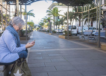 Side view of senior woman using phone while sitting by walkway