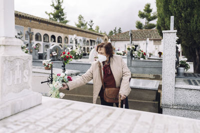 Senior woman wearing face mask putting flowers on grave in cemetery