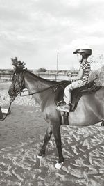 Girl sitting on horse at sand against cloudy sky