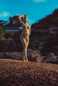 A pregnant girl in gold glitter stands on a coral stone in the middle of the ocean