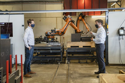 Businessmen wearing face mask standing with social distance against robotic machine at factory