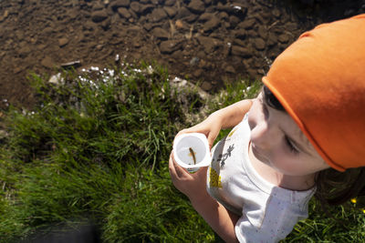 Cute little girl holding disposable cup with fish inside it standing on lakeshore