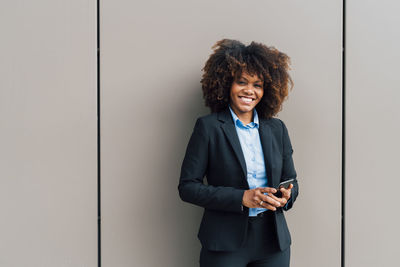Happy afro businesswoman holding mobile phone standing in front of wall