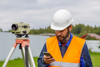 A civil engineer with an optical level looks into a mobile phone. a bearded man is looking 