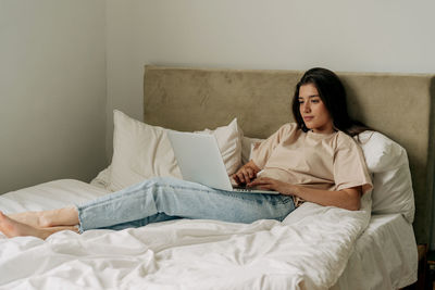 Young brunette freelancer in t-shirt and jeans lying on bed working on laptop