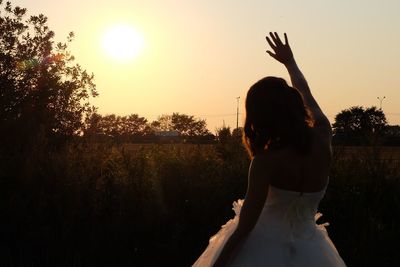 Rear view of bride waving while standing by plants against sky during sunset