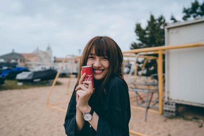 Portrait of smiling young woman holding camera while standing outdoors