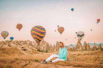 Thoughtful mid adult man sitting on field against hot air balloons flying in mid-air
