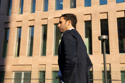 From below side view of thoughtful male in formal clothes looking away with pensive face while standing on sunny street near building with shadow on face